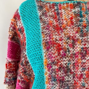 May include: A colorful, handmade crochet sweater with a unique texture. The sweater features a vibrant mix of red, orange, purple, and white yarns. A bright turquoise panel runs down the front, adding a striking contrast to the design. The sleeves have a pink accent.