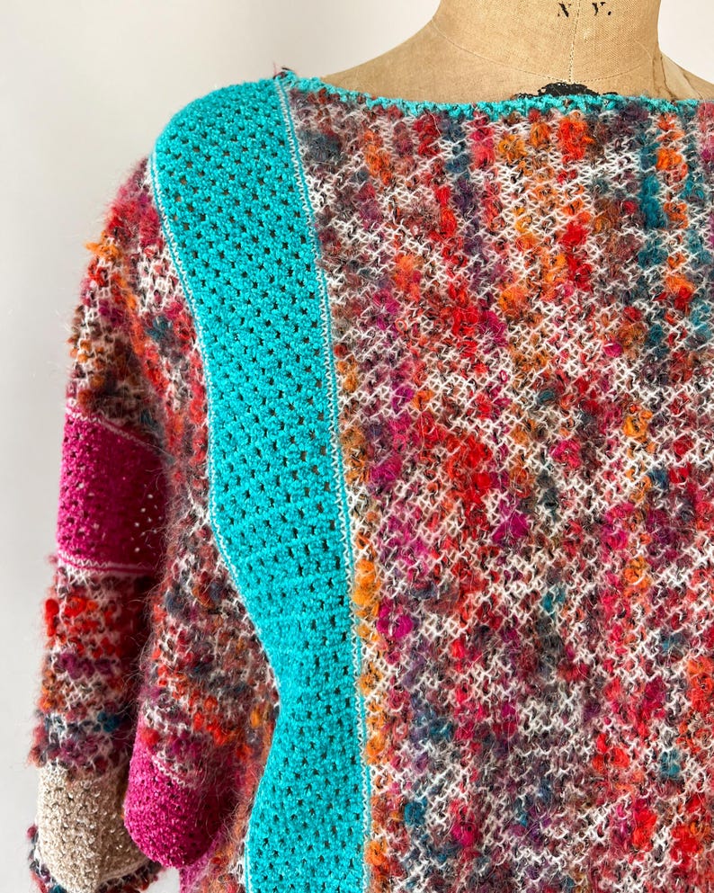 May include: A colorful, handmade crochet sweater with a unique texture. The sweater features a vibrant mix of red, orange, purple, and white yarns. A bright turquoise panel runs down the front, adding a striking contrast to the design. The sleeves have a pink accent.