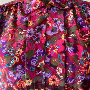 May include: A vintage floral print blouse featuring a vibrant pattern of pink, purple, orange, and teal flowers on a burgundy background. The blouse has a collared neckline, a single brown button, and pleated detailing down the front.
