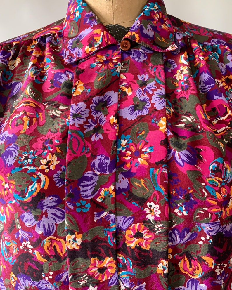 May include: A vintage floral print blouse featuring a vibrant pattern of pink, purple, orange, and teal flowers on a burgundy background. The blouse has a collared neckline, a single brown button, and pleated detailing down the front.