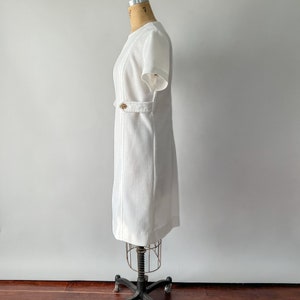 May include: A white, short-sleeved dress with a textured fabric and a subtle pattern. The dress features a decorative gold-colored button detail at the waist and is displayed on a vintage dress form. The dress has a classic, tailored silhouette.