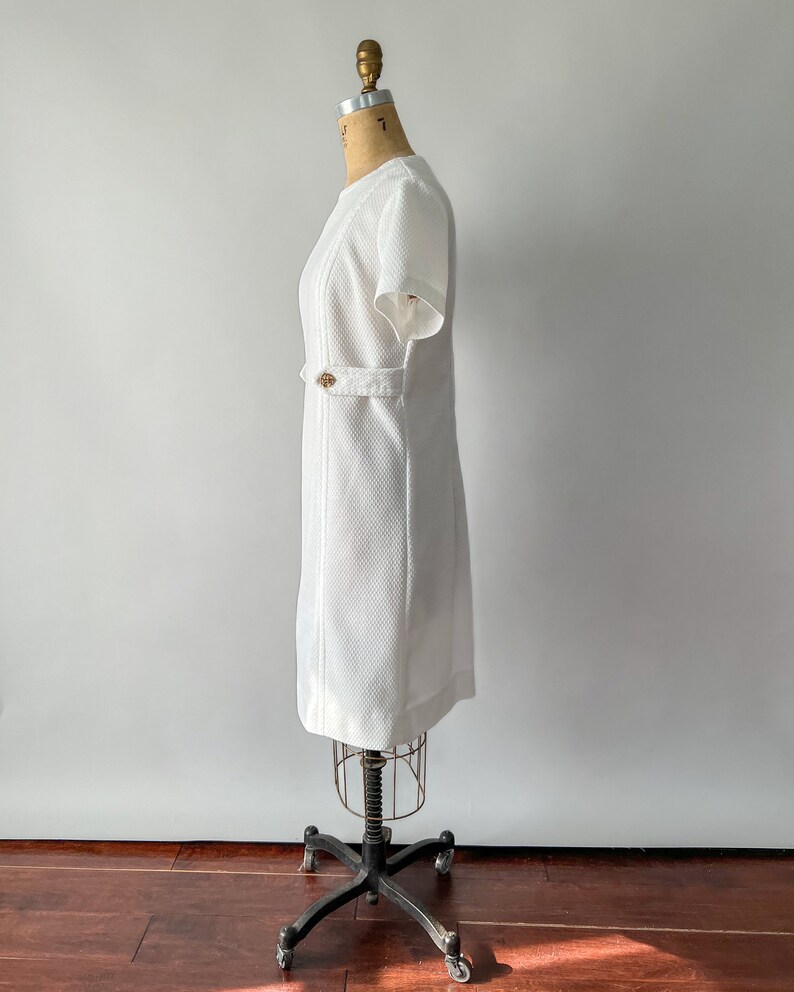 May include: A white, short-sleeved dress with a textured fabric and a subtle pattern. The dress features a decorative gold-colored button detail at the waist and is displayed on a vintage dress form. The dress has a classic, tailored silhouette.