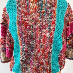 May include: A colorful, textured crochet sweater with a boat neck. The sweater features a mix of vibrant colors, including red, orange, pink, and teal. Teal crochet panels run down the front and back, and the sleeves have a patchwork design.