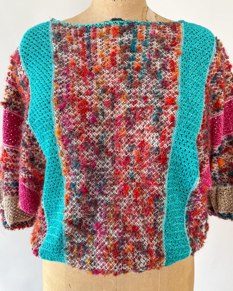 May include: A colorful, textured crochet sweater with a boat neck. The sweater features a mix of vibrant colors, including red, orange, pink, and teal. Teal crochet panels run down the front and back, and the sleeves have a patchwork design.