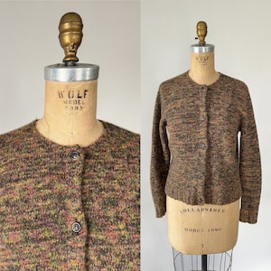 May include: A vintage button-down cardigan sweater in a speckled pattern of brown, green, yellow, and orange. The sweater is displayed on a vintage Wolf Model Form mannequin. The sweater has a round neckline and long sleeves.