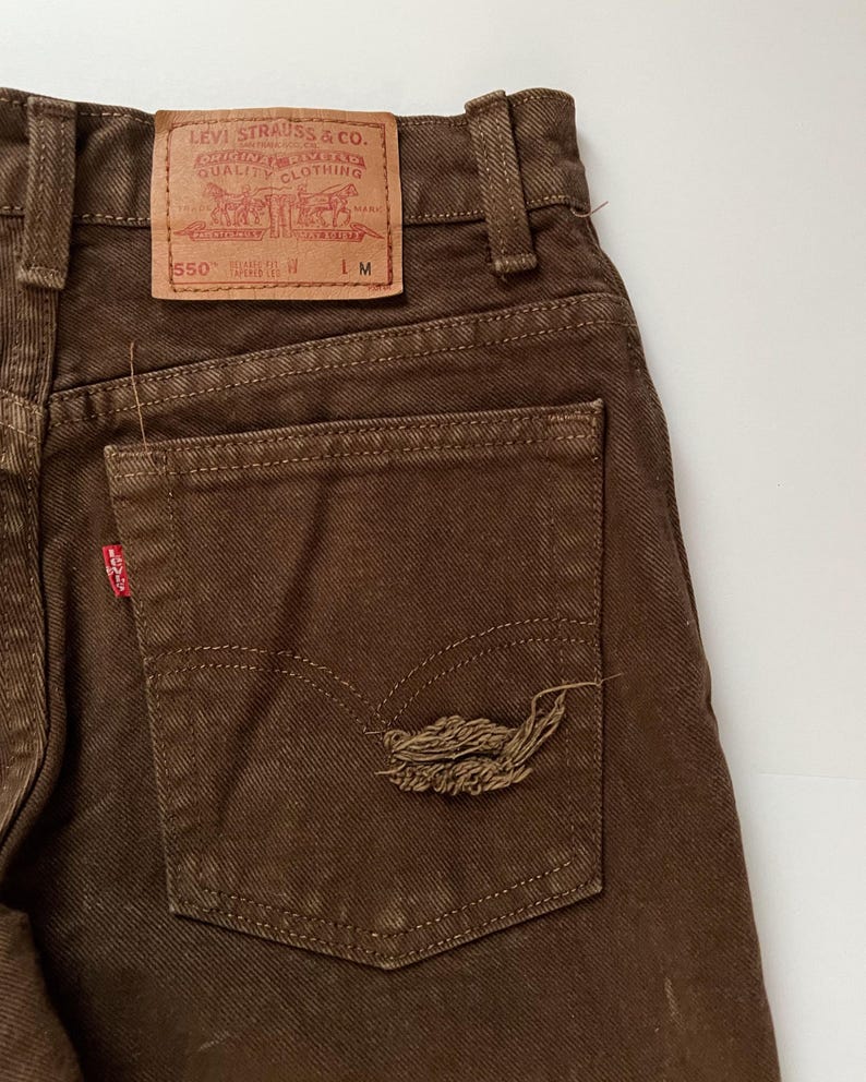 Puede incluir: Vaqueros de mezclilla marr&oacute;n con un parche de Levi Strauss & Co. en el bolsillo trasero. El parche tiene el texto "Levi Strauss & Co.  San Francisco, Cal.  Original  Quality Clothing  Trade Mark  Registered  550" y "L.M.".