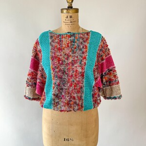 May include: A colorful, textured sweater with a round neckline and short sleeves. The sweater features a vibrant mix of red, orange, pink, and teal yarns. Teal panels run down the front and back, and the sleeves have pink and beige accents. The sweater is displayed on a vintage dress form.