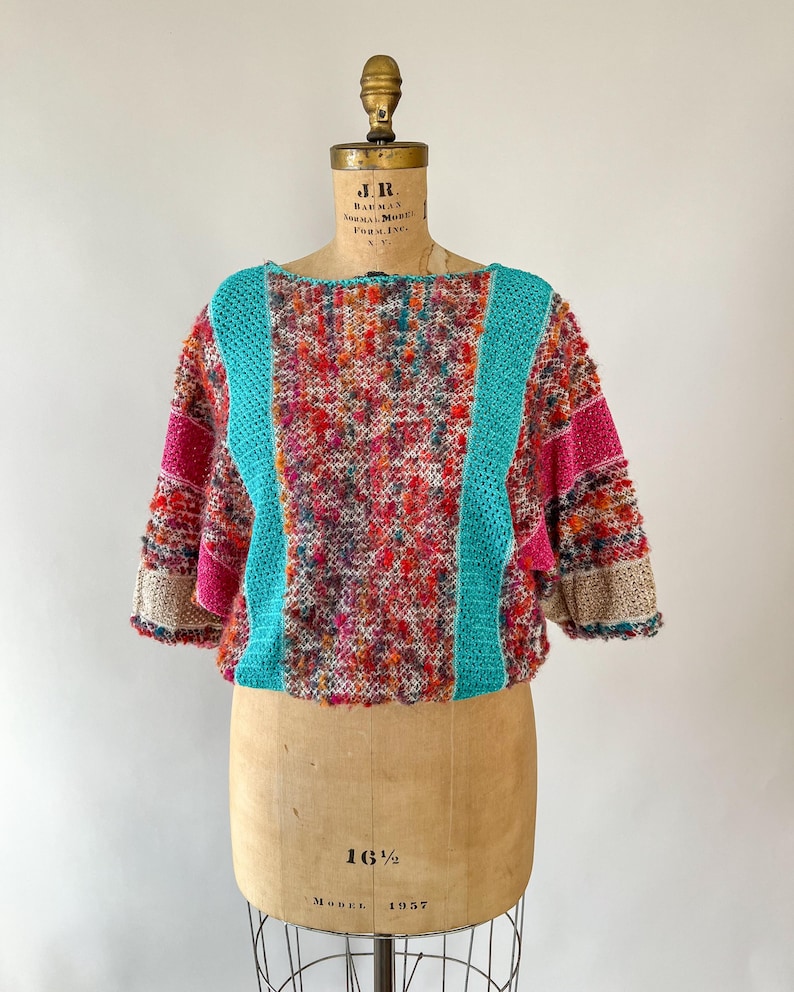 May include: A colorful, textured sweater with a round neckline and short sleeves. The sweater features a vibrant mix of red, orange, pink, and teal yarns. Teal panels run down the front and back, and the sleeves have pink and beige accents. The sweater is displayed on a vintage dress form.