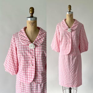 May include: A vintage pink and white gingham two-piece suit. The jacket has a rounded collar and short sleeves. The skirt is knee-length. The suit is displayed on a mannequin. The jacket has three buttons and a decorative white brooch.