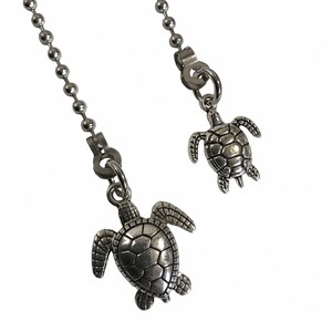 May include: Two silver chain pulls with turtle charms. The larger turtle charm has a detailed shell and is hanging from a longer chain. The smaller turtle charm has a simpler shell and is hanging from a shorter chain.