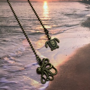 May include: Two bronze fan pull chains with a sea turtle and an octopus design. The chains are against a background of a beach with waves and a sunset.