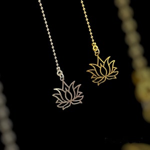 May include: Two lotus flower pendants, one silver and one gold, hang from ball chains against a black background. The pendants are detailed with openwork petals, showcasing a delicate floral design. The chains are suspended, creating a sense of floating elegance.