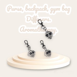 May include: Three silver heart-shaped essential oil diffusers with black metal grates. The diffusers are attached to key rings and can be hung on a purse, backpack, or gym bag. The text "Purse, backpack, gym bag Diffusers. Aromatherapy." is written in white on a white background.