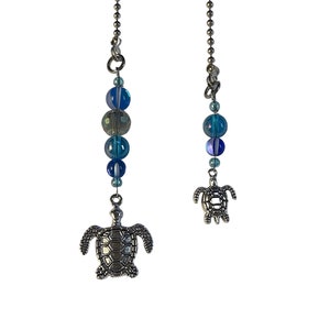 May include: Two silver turtle charms hang from a silver chain. The top charm has a string of blue and white iridescent beads.