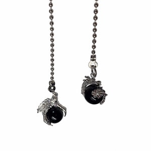 May include: Two decorative ceiling fan pull chains. Each chain has a silver-colored metal ball chain with a decorative charm. One charm is a dragon claw holding a black sphere, and the other is a dragon wrapped around a black sphere.