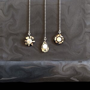 May include: Three silver chains with decorative pendants hanging from a dark gray surface. The pendants are made of black and white shell with a silver trim.