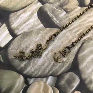 May include: A bronze chain with a mermaid and anchor charm. The chain is on a background of gray and brown stones.