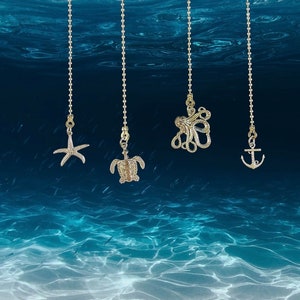 May include: Four gold-toned light pull chains with different sea-themed charms: a starfish, a turtle, an octopus, and an anchor. The chains are hanging against a blue background that resembles water.