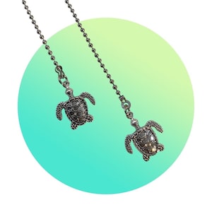 May include: Two silver turtle-shaped fan pull chains hanging against a green and blue gradient background.