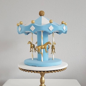 May include: A carousel cake topper with a light blue and white canopy, gold accents, and gold horse figurines. The topper sits on a white and gold cake stand with a gold base. A decorative piece for a cake.
