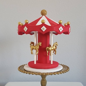 May include: A red and white carousel cake topper with gold accents. The topper features gold horses, a red base, and a striped canopy. It is displayed on a gold pedestal cake stand, perfect for a birthday or special event.