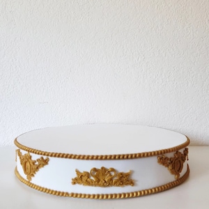 May include: A white, round cake stand with gold decorative trim and ornate gold embellishments. The stand has a flat top and a decorative border with a rope-like design. The gold accents provide a regal and elegant appearance.