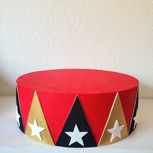May include: A red, round circus-themed cake stand. The stand features alternating black, gold, and white star-adorned triangles around the base. The top is a solid red color, creating a vibrant and festive design.