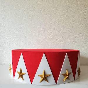 May include: A red, cylindrical decorative piece with white triangles and gold stars. The design is reminiscent of a circus tent or a festive celebration. The object is set against a plain white background.