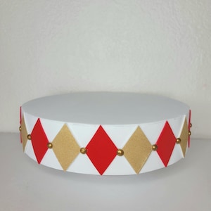 May include: A white, round cake stand with a decorative band of red and gold diamond shapes. Small gold beads accent the diamonds. The stand is designed to hold cakes or other desserts.