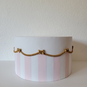May include: A round, white box with vertical pink stripes. The box is decorated with a gold-colored rope and bow design around the top. The box is sitting on a white surface.