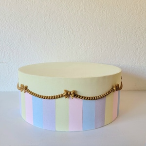 May include: A pastel-colored round box with vertical stripes in light blue, lavender, and yellow. The box is decorated with a gold-colored rope and bow design around the middle. The top of the box is a solid pale yellow.
