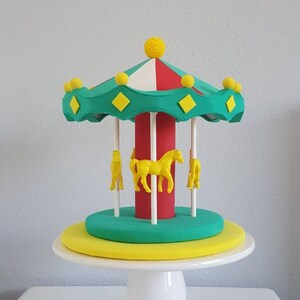 May include: A colorful carousel cake topper with a white base. The carousel features a green and red canopy with yellow accents, a red center pole, and yellow horse figurines. The cake sits on a white pedestal.