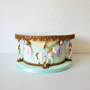May include: A carousel-themed cake with a light green base and a gold-colored decorative trim. The cake features white carousel horses with gold manes and blue saddles, along with pastel-colored fabric accents. The cake sits on a light green base with a matching ribbon.