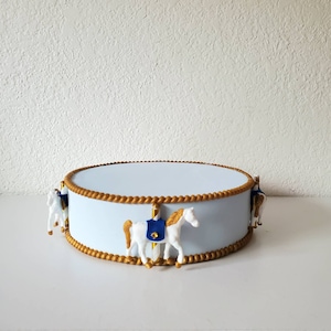May include: A light blue carousel-themed cake stand with gold trim and decorative white horses. The stand has a round shape and is designed to display cakes or other treats. The horses have gold manes and blue saddle blankets.