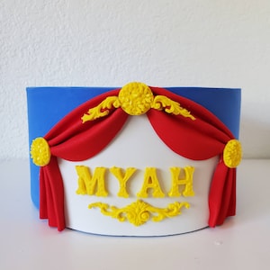 May include: A blue and white decorative box with red curtains and yellow embellishments. The name "MYAH" is displayed in yellow letters. The box has a circus or theater theme.