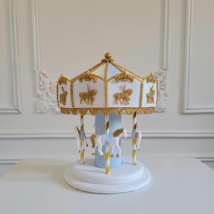May include: A miniature carousel with a white and gold roof, blue and gold poles, and white horses. The carousel has a white base and is decorated with gold accents. The horses have brown manes and tails.