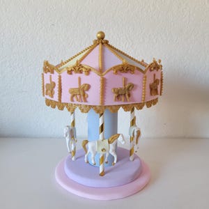May include: A miniature carousel with a pink and gold roof, featuring gold horse figures and white horses. The carousel has a light blue center column and sits on a lavender and pink base. Decorative and whimsical.