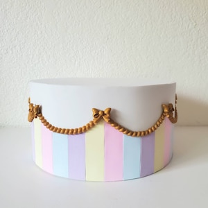 May include: A round, pastel-colored cake stand with vertical stripes in pink, blue, yellow, and lavender. The top is white, and a gold-colored garland with bows decorates the edge. The stand is on a white surface.