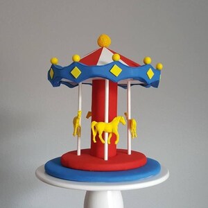 May include: A colorful carousel cake with a red and white striped roof, blue trim with yellow diamond accents, and yellow horse figurines. The cake sits on a white pedestal.
