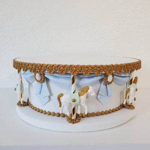 May include: A white carousel cake stand with gold accents and light blue ribbons. The stand features white carousel horses and a decorative gold trim around the top and bottom. The cake stand is round and sits on a white base.