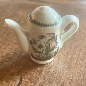 Puede incluir: Una pequeña tetera de cerámica blanca con un diseño floral. La tetera tiene un borde verde y un patrón floral con flores rosas y blancas.