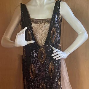 Puede incluir: Vestido negro con lentejuelas y detalles dorados en forma de hoja. El vestido presenta un escote en V profundo y una tela beige transparente en capas en el lateral. El vestido se exhibe en un maniquí.