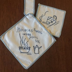 May include: Three white cotton pot holders with blue trim. One pot holder has a handle and says "Holders Three Inside You'll See" with an embroidered image of a pot with steam coming out of it and a teapot. The other two pot holders have embroidered images of teapots.
