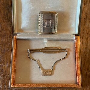 Puede incluir: Un clip de corbata de oro con una cadena y un pequeño amuleto rectangular en una caja forrada de terciopelo. El amuleto tiene un diseño grabado.