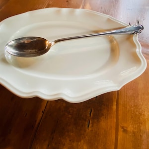 May include: An off-white, oval-shaped serving platter with a scalloped edge, paired with a silver spoon. The spoon rests on the platter, showcasing a classic design. The spoon's length is approximately 8 inches.