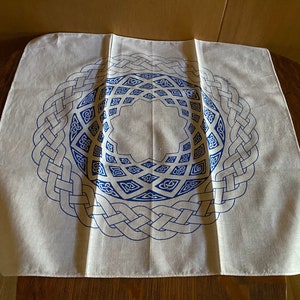 May include: White cotton square cloth with a blue Celtic knot design. The knot design is a circle with a repeating pattern of interlacing lines.
