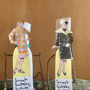 May include: Two vintage paper dolls, each mounted on a stand. One doll wears a geometric patterned coat, the other a black coat with a white collar. Each has a yellow tag that reads "Great Gatsby 8-14-99".