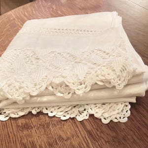 May include: A stack of folded white linens with intricate lace detailing along the edges. The linens are neatly arranged, showcasing the delicate patterns and textures of the lace. The linens are on a wooden surface.