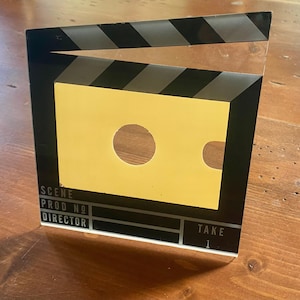 May include: A black and yellow film clapperboard with the text "SCENE", "PROD NO", "DIRECTOR", and "TAKE 1". The clapperboard has a yellow center with a circular and a curved cutout.
