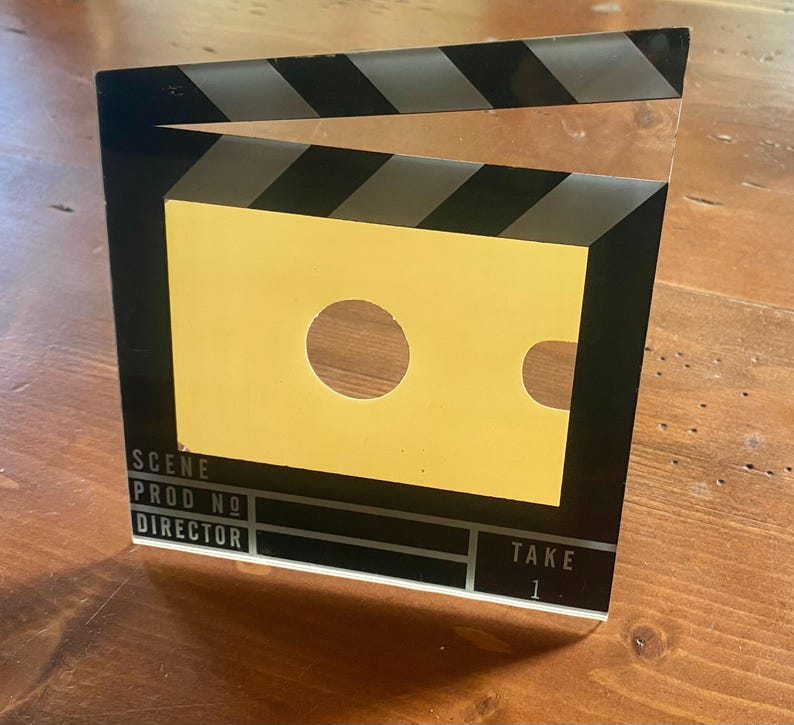 May include: A black and yellow film clapperboard with the text "SCENE", "PROD NO", "DIRECTOR", and "TAKE 1". The clapperboard has a yellow center with a circular and a curved cutout.